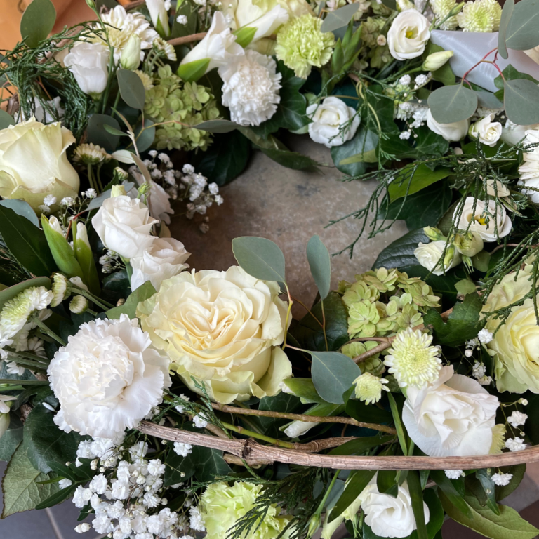 Kranz für die Urne mit weißen und grünen Blumen wie Rosen und Nelken, umgeben von Eukalyptus und zarten Pflanzen.