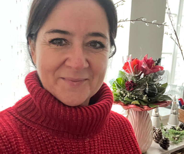 Jacqueline Turowsky privat mit dunklen Haaren trägt einen roten Pullover, im Hintergrund ein Blumenarrangement.